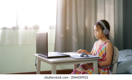 Asiatische Kinderstudentin oder Kindermädchen, die auf einem Computer-Notebook lernen, ein Headset zu tragen, um online per Videoanruf zu lernen, oder eine Person, die von zu Hause aus auf dem Laptop lernt, indem sie zurück zur Schule auf Fenster mit warmem Licht kommt