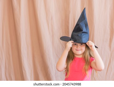 Beautiful cute girl 6-7 years old with long blond hair in a red t-shirt holds on to the witch's hat celebrating halloween on an isolated background. Concept: october holiday halloween all saints day 