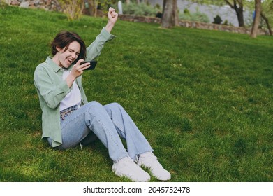 Jonge vrouw in groene spijkerbroek zit op gras in het lentepark van de stad buiten spelen racen op mobiele mobiele telefoon houd gadget smartphone pc-videogames vast, doe winnaargebaar. Mensen stedelijk levensstijlconcept.