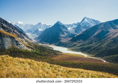 Wonderful alpine landscape with mountain lake and mountain river in valley with forest in autumn colors on background of snowy mountains silhouettes under blue sky. Beautiful mountain valley in autumn