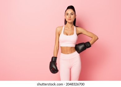 strong woman in sportswear and boxing gloves standing with hand on hip on pink