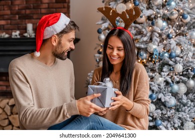 自宅のクリスマスツリーの横に座っている彼の女性の妻のガールフレンドにクリスマスプレゼントボックスを贈る男性の夫のボーイフレンドの配偶者。あけましておめでとう！