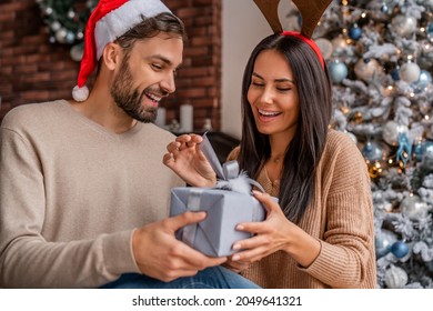 自宅のクリスマスツリーの横に座っている彼の女性の妻のガールフレンドにクリスマスギフトボックスのプレゼントを与える男性の夫のボーイフレンドの配偶者。あけましておめでとう！