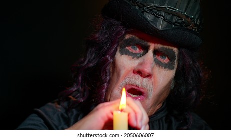 Creepy mature man grandfather with Halloween stylish witcher makeup. Old scary grandpa making voodoo magic rituals with candle and smiles terribly. Thematic party. Sinister sorcerer senior guy vampire