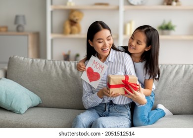 Xin chúc mừng. Con gái châu Á yêu thương chúc mẹ ngạc nhiên nhân Ngày của Mẹ, tặng mẹ hộp quà và thiệp thủ công, ngồi trên ghế sofa trong nhà, không gian trống