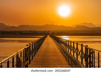 エジプトの砂漠の山々のシルエットに絵のように美しい明るいオレンジ色の夕日と海岸線が海の水に反射し、休暇中の夏の夜に日光につながる木製の橋の道