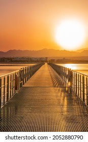 Wooden and metal bridge road or pontoon leading to bright picturesque orange sunset over desert mountains of Egypt with coastline and reflections in sea water at sunny vacation evening. Vertical image
