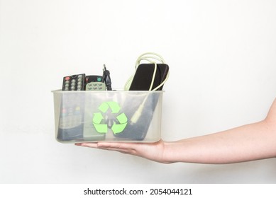 Una mano que sostiene una caja de plástico transparente con el símbolo de reciclaje que contiene controles remotos de televisión, un teléfono y otros dispositivos electrónicos en un fondo blanco