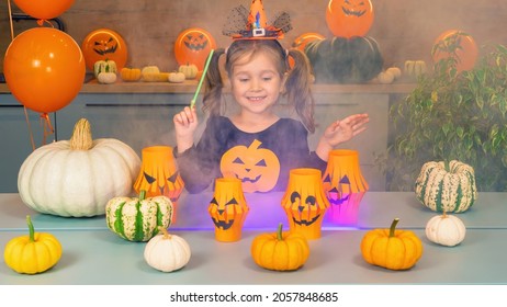Cheerful little girl shows magic tricks with a magic wand for Halloween. Multi-colored smoke over the table of the sorceress. The little witch has bewitched the pumpkins. Fun games for Halloween.