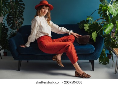秋のファッションのコンセプト: オレンジ色の帽子、ビンテージ スタイルのブラウス、トレンディなキュロット、ヒョウ柄のローファー シューズを身に着けているエレガントな女性は、青いインテリアでソファでポーズします。コピー、テキスト用の空白