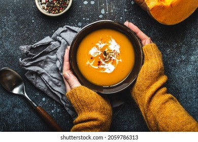 Tangan wanita dengan sweter rajutan kuning memegang mangkuk dengan sup krim labu di latar belakang batu gelap dengan sendok dihiasi dengan potongan labu segar, pemandangan atas. Konsep makan malam musim gugur yang nyaman