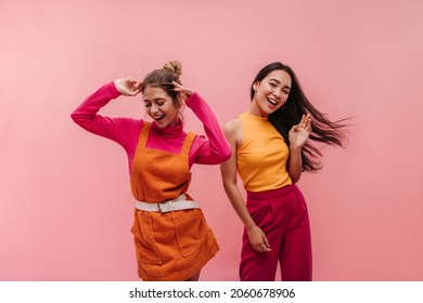 Energetic young two interracial girls dancing side by side and waving hands in studio. Models with light brown and black hair in light spring orange and pink clothes move to music with smile on face.