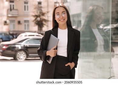Mujer asiática joven de negocios morena con una computadora portátil en sus manos que no está en pleno crecimiento sonriendo a la cámara. La dama con anteojos se para fuera del edificio de oficinas durante su hora de almuerzo.