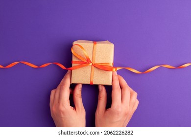 Flat lay of brown gift box with a orange satin ribbon bow and hands on it on vibrant purple background with copy space. Holiday autumn and Halloween concept.