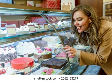 Een mooie jonge vrouw kijkt naar een glazen vitrine met snoep. Lachende blondine in een beige jas kiest een taart.