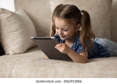 Szczęśliwa urocza mała dziewczynka 6s leżąca na wygodnej kanapie z gadżetem touchpada w rękach, zaangażowana w granie w gry wideo, oglądanie rozrywkowych treści w sieciach społecznościowych, spędzanie czasu online.