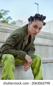 Cool teenage girl with stylish hairstyle makeup sits on steps outdoors wears fashionable green clothes earrings and rings looks self confident at camera belongs to youth subculture. Vertical shot
