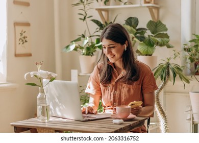 Mulher jovem e otimista usando um laptop e um smartphone em casa. Interior aconchegante com plantas. Conceito de trabalho remoto, negócios, freelancer, compras online, ensino a distância e selva urbana. Foco seletivo.
