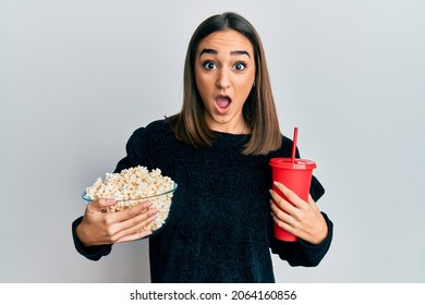 Young brunette girl eating popcorn and drinking soda afraid and shocked with surprise and amazed expression, fear and excited face. 