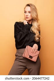 Young beautiful blond woman in a black blouse and brown pants holds a pink handbag and posing on a beige background. Fashion style.