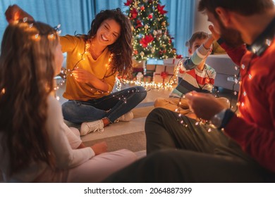 Parents and children having fun spending Christmas Eve together at home, sitting on the floor by the Christmas tree, playing with Christmas lights