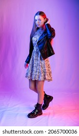 potret panjang penuh seorang gadis. Model dengan sepatu bot kasar, gaun sifon tipis, dan jaket kulit. latar belakang berwarna. cahaya biru, merah, ungu pada latar belakang dan orang