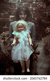 Een angstaanjagende spookvrouw met enge, vervaagde ogen die sinister lacht met een babypop tussen verdord gebladerte bij een verwoeste muur. Horrorfilm, thriller. Halloween.