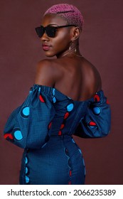 Portrait of a bald African woman with her back to the camera, looking to the left side, showing the African costumes. She has dyed her hair pink and is wearing sunglasses on a brown background.