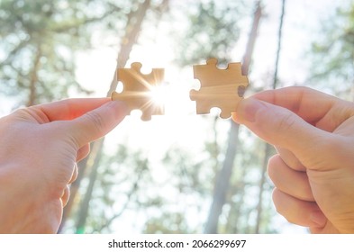 Jigsaw puzzle against a forest background. Two hands connecting two puzzle pieces with sunlight and rays coming through. Teamwork and business cooperation concept.