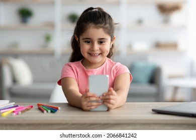 Gadgets And Kids. Cute Little Arab Girl Using Smartphone At Home, Adorable Female Child Playing Mobile Games Or Watching Cartoons Online On Cellphone While Sitting At Table In Living Room, Closeup
