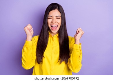茶色の髪型の写真は、若い女性の手の拳を祝うために目を閉じて叫び、紫色の背景に黄色のシャツを着用します