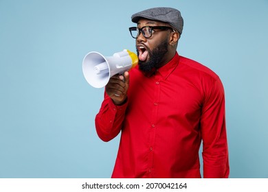Shocked amazed young bearded african american man 20s wearing casual red shirt eyeglasses cap standing screaming in megaphone looking aside isolated on pastel blue color background studio portrait