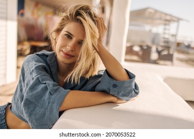 手で彼女の頭を保持しながら優しくカメラに探している美しい若い女性のクローズ アップ。ブルーのルーズなシャツを着た片側に乱れた髪のブロンド。朝の楽しい時間のコンセプト
