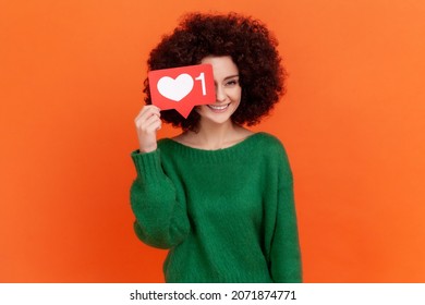 Feliz mujer adulta joven positiva con peinado afro usando suéter verde de estilo casual cubriendo su ojo con el corazón de las redes sociales como icono. Disparo de estudio interior aislado sobre fondo naranja.