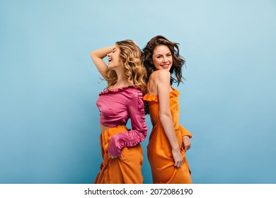 青い色のスタジオ ショットで元気に笑って背中合わせに 2 つのかわいい白人の女の子が立っています。ブルネットは裸の肩を持つオレンジ色のドレスを着ています。ピンクのブラウスとオレンジのズボンの金髪。