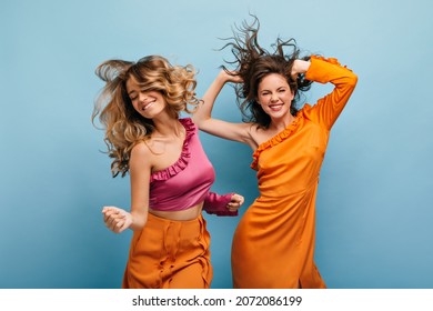 Cheerful showy caucasian young women fooling around on isolated blue background. Their wavy, loose hair flies in all directions. Girls are dressed in bright summer outfits.