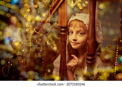 Portrait of an amusing little girl in a nightgown and nightcap sitting joyfully on a wooden staircase festively decorated on Christmas. Christmas magic and dreams.