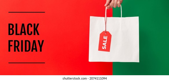 ブラック フライデーのバナー。赤と緑の背景に販売価格タグ付きの白い白紙の買い物袋を持っている女性の手。販売、割引、ショッピングのコンセプト
