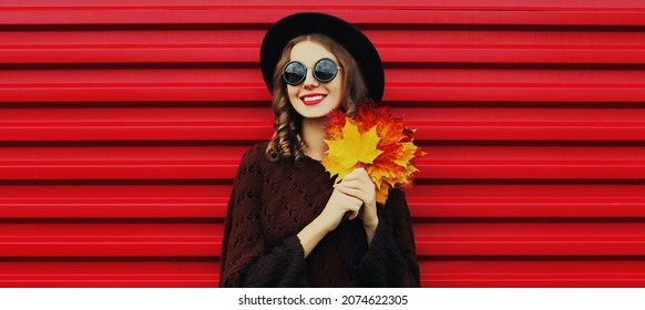 Portrait of beautiful young woman with yellow maple leaves wearing a brown knitted poncho, black round hat on red background