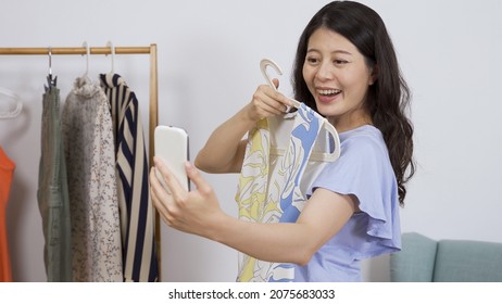 potret wanita memegang telepon sedang mencoba pakaian dan menunjukkan bagian belakang one piece sambil meminta saran temannya selama panggilan video.