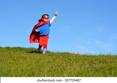 Niña superhéroe (niña) corre en un campo verde contra un fondo de cielo azul dramático con espacio de copia. Foto conceptual de superhéroe, poder femenino, juego de simulación, infancia, imaginación.
