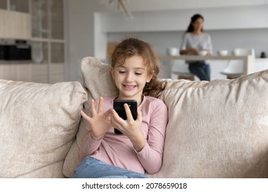 Smiling little girl having fun with smartphone while mother cooking on background, cute kid looking at phone screen, playing mobile device game, watching cartoons, sitting on cozy couch at home