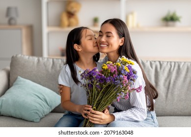 Con yêu mẹ. Cô gái châu Á xinh đẹp hôn má mẹ và tặng bó hoa, chúc mừng mẹ nhân ngày lễ, ngày phụ nữ hoặc sinh nhật của mẹ, ngồi trên ghế sofa