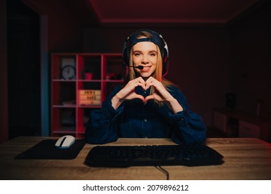 Positive female gamer in a headset sits at night in a room with a red light, plays computer games and streams, looks at the camera with a smile on her face and shows a heart gesture.