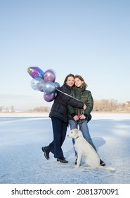 dos mujeres con perro blanco y un montón de globos. paseo divertido en invierno en la nieve. una hija adulta sonriente de entre 17 y 18 años y una madre de unos 40 años miran la cámara, un tierno momento familiar
