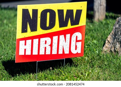 Sign text closeup for help wanted now hiring in English with red and yellow colors as signpost on grass for store shop business during corona virus covid 19 pandemic