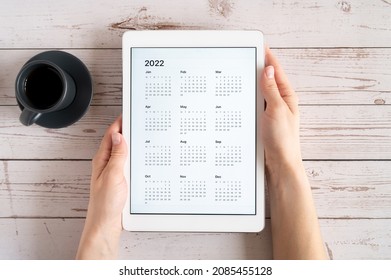 máy tính bảng với ứng dụng mở lịch cho năm 2022 và tách trà hoặc cà phê trên tay người phụ nữ trên nền gỗ. khái niệm kinh doanh hoặc để thực hiện các mục tiêu danh sách với việc sử dụng công nghệ. nhìn từ trên xuống, nằm phẳng