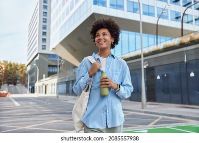 カジュアルなシャツを着た幸せな巻き毛の若い女性のウエストアップショットは、生地のバッグを持ち、ボトルの笑顔に新鮮な水を入れ、ガラスの高層ビルの間で街を積極的に散歩し、余暇を楽しんでいます。