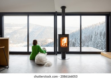 Frau sitzt mit Hund in der Nähe von Kamin und Panoramafenster im modernen Wohnzimmer mit herrlichem Blick auf schneebedeckte Berge. Konzept der Erholung in Häusern oder Hütten in der Natur. Idee der Flucht aus dem Alltag
