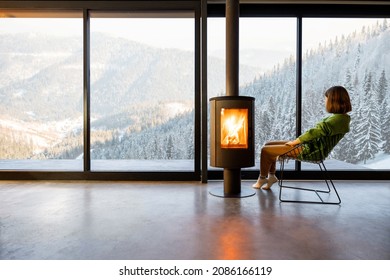 Wanita duduk di dekat perapian di ruang tamu modern dengan pemandangan pegunungan bersalju. Konsep istirahat di rumah atau kabin di alam. Kesendirian di alam dan melarikan diri dari kehidupan sehari-hari